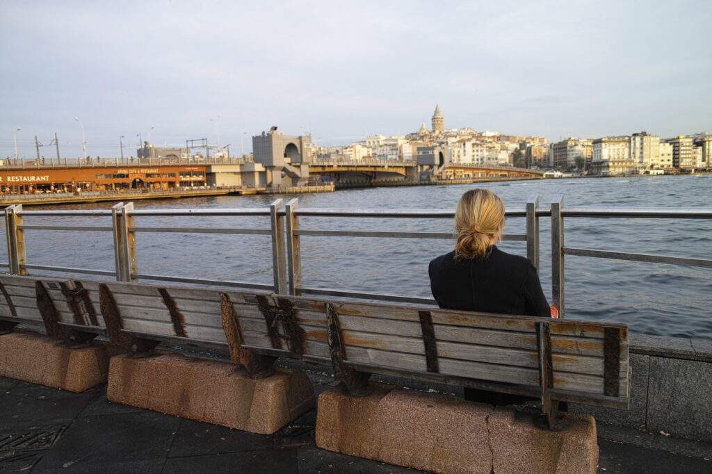A picture of a woman sitting on a bench looking at the sea with a city at the background